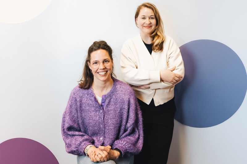 Pia Brand sitzt vor einer hellen Wand, auf der große runde Farbflächen zu sehen sind, rechts neben ihr steht Andrea Ruppert, beide lächeln in die Kamera.