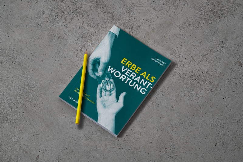 Das Buch "Erbe als Verantwortung" liegt auf einem Betonboden, darauf ein gelber Bleistift. Auf dem Buchcover sieht man eine ältere Hand, die einen Kompass in eine jüngere Hand legt.